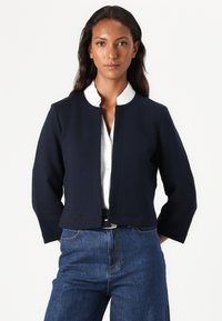 Marineblauw cropped jasje met een ronde halslijn, lange mouwen en een gestructureerde stof. Gepaarde met een witte blouse met kraag en denim jeans.