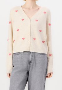 Ljusbeige kardigan med V-ringning och knäppning. Har små rosa hjärtan över hela. Tillverkad av mjukt, texturerat tyg med ribbade ärmslut.