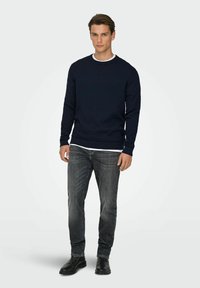 Pull texturé bleu marine avec poignets et ourlet côtelés, porté par-dessus une chemise blanche, associé à un jean gris et des bottes noires.