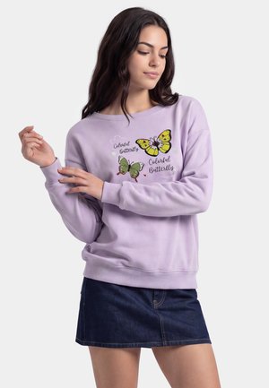 Kvinde iført en lavendel sweatshirt med gule og grønne sommerfugleprint og tekst, kombineret med et mørkt denimskørt, poserer med hævede hænder.