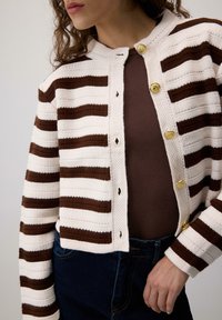 Femeie îmbrăcată într-un cardig tricotat cu dungi albe și maro, cu butoane aurii, peste un top maro și blugi de culoare albastră închisă, stând în fața unui fundal simplu.