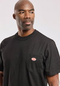 Schwarzes Baumwoll-T-Shirt mit kurzen Ärmeln, ausgestattet mit einer linken Brusttasche und einem roten und weißen Logopatch. Glatte Textur, klassischer Schnitt.