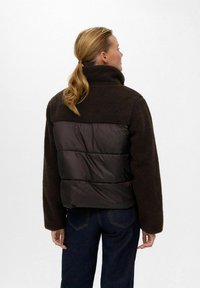 Frau mit blondem Pferdeschwanz trägt eine braune Fleecejacke mit gestepptem Rückenteil und dunkelblaue Jeans, blickt von der Kamera weg.