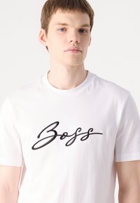 Fehér pamut póló, amelyen egy kiemelkedő fekete "Boss" logó található a mellkason. Egyszerű, kerek nyakú dizájn.