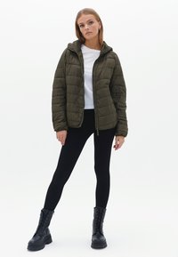 Olijfgroene gewatteerde jas met een capuchon, ritssluiting, horizontale quilting en lange mouwen, gecombineerd met zwarte leggings en combatboots.