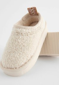 Beige toffel med fluffig, texturerad ovandel, vadderad krage och gummisula. Har en vävd tygöglä vid hälen för enkel påtagning.
