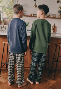 Due ragazzi stanno schiena contro schiena, indossando maglie a maniche lunghe: una blu scuro, l'altra verde. Portano pantaloni del pigiama a quadri, grigi e blu scuro/verde, con orli elastici.