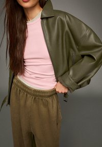 Eine olivgrünen Lederjacke, eine hellrosa Bluse und lockere olivgrüne Hose mit elastischem Bund tragend, mit der Hand nah an der Hüfte.