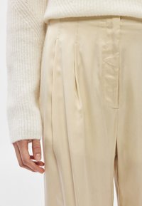 Χέρι ακουμπισμένο στο πλευρό, φορώντας ένα κρεμ πλεκτό πουλόβερ και μπεζ σατέν παντελόνι με πιέτες και ψηλή μέση, σε ουδέτερο φόντο.