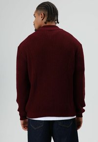 Man met gevlochten haar, van achteren gezien, draagt een donkerrode grofgebreide trui over een wit overhemd en donkerblauwe spijkerbroek.