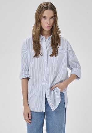 Joven con cabello largo y castaño, vistiendo una camisa blanca holgada con finas rayas negras y jeans azul claro, con la mano en el bolsillo, fondo neutro.