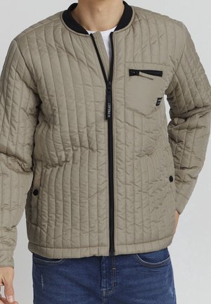 Beige quilted bomber jacket with a black ribbed collar, front zip closure, and a zippered pocket on the left chest. Features stitched patterns.