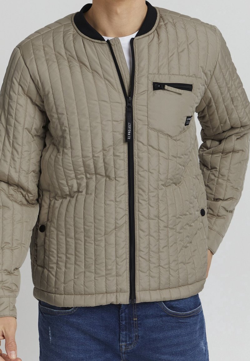 Beige quilted bomber jacket with a black ribbed collar, front zip closure, and a zippered pocket on the left chest. Features stitched patterns.