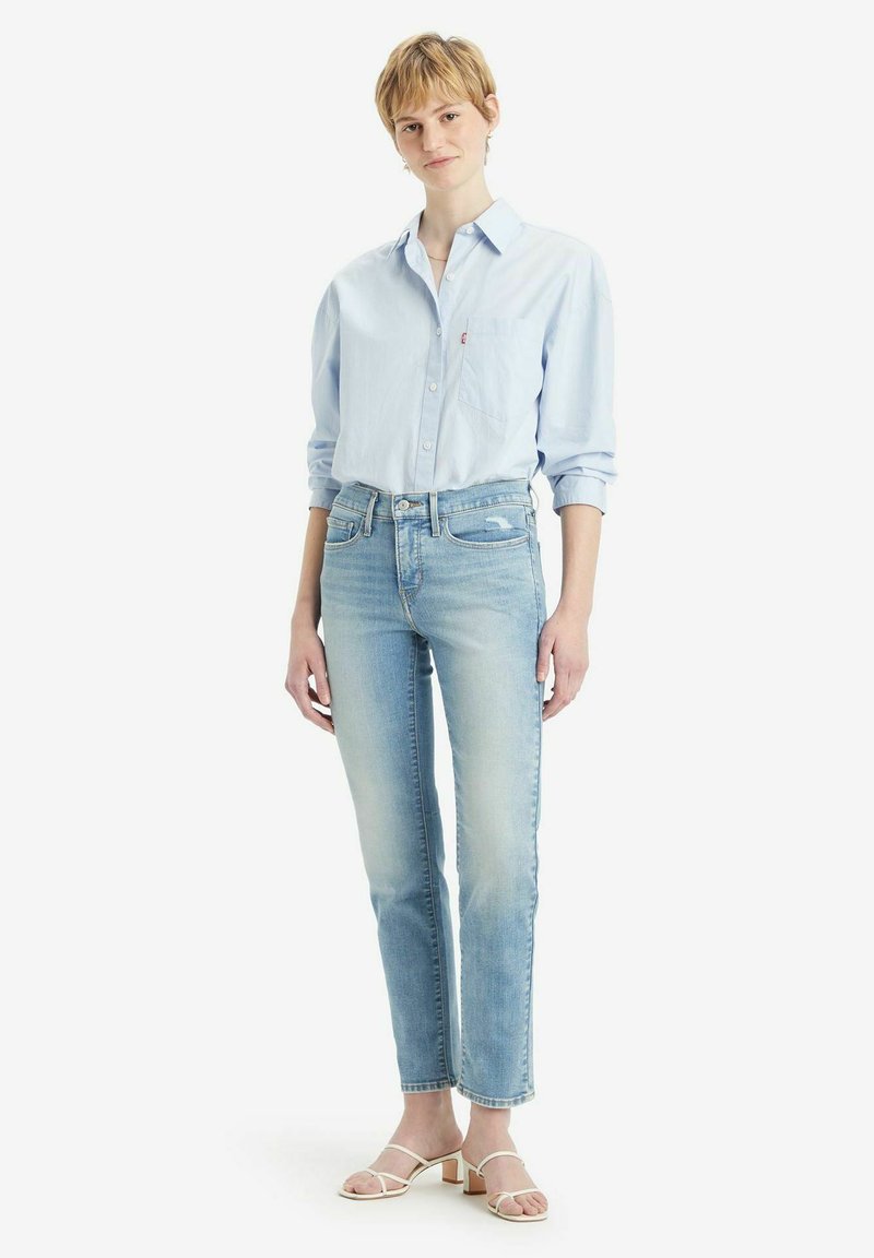 Camisa de botones azul clara con un bolsillo en el pecho, combinada con jeans rectos azul claros y sandalias de tacón blancas con tiras. Diseño simple.