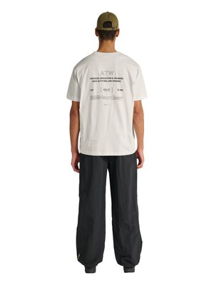 Man wearing olive green cap, white T-shirt with black text on back, and loose black pants, standing facing away on white background.