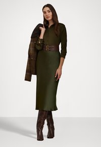 Lang, olivengrønn kjole med en tettsittende silhuett og et bredt, brunt belte. Modellen har på seg en mørk rutete blazer over den ene skulderen og brune hælboots.