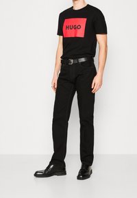 Schwarzes Baumwoll-T-Shirt mit einem roten, rechteckigen Logo mit der Aufschrift "HUGO." Kombiniert mit schwarzer Jeans und schwarzen, polierten Schuhen. Einfaches Design, figurbetonte Form.