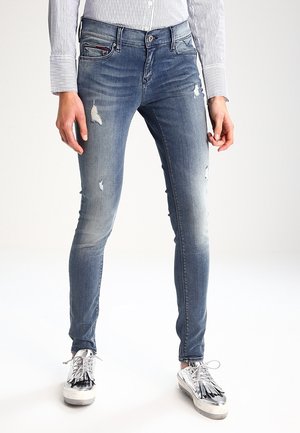 Femme portant un jean skinny bleu délavé avec des empiècements usés et des baskets métalliques argentées, debout devant un fond blanc uni.