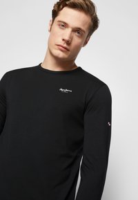 Schwarzes langärmliges T-Shirt aus glattem Stoff. Verfügt über ein kleines weißes Logo auf der Brust und einen kontrastierenden gestreiften Akzent am Ärmel.