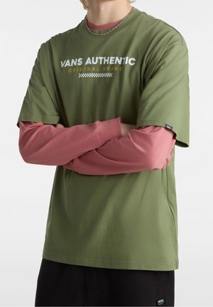 Person iført olivengrøn Vans Authentic t-shirt over langærmet lyserød skjorte med korslagte arme, sorte bukser og en sølvkæde.