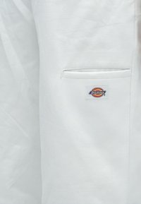 Λευκό ύφασμα σε κοντινή λήψη με μια σχισμή τσέπης και ένα ορθογώνιο λογότ υπο Dickies ραμμένο κάτω από το άνοιγμα της τσέπης.