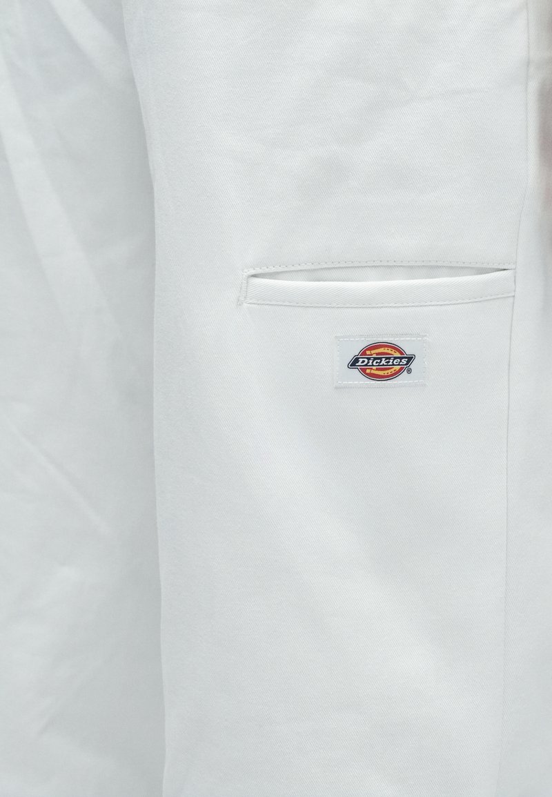 Primo piano di un tessuto bianco con una tasca a fessura e una toppa rettangolare con il logo Dickies cucita sotto l'apertura della tasca.