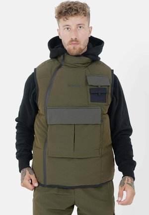 Gilet verde imbottito con colletto alto, numerose tasche e chiusura con zip. Indossato sopra una maglietta nera a maniche lunghe, caratterizzata da un tessuto testurizzato.