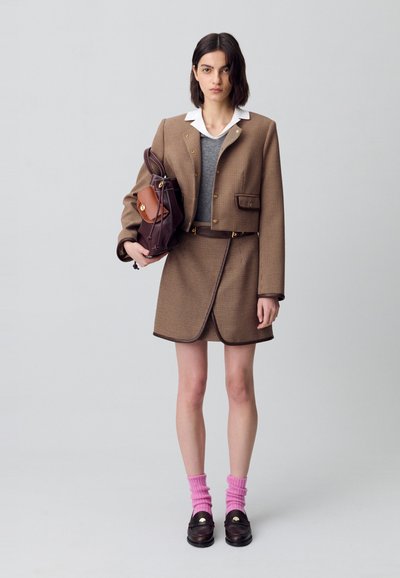 Modella in un blazer a quadri sopra un top grigio e una gonna, che tiene una borsa marrone, indossando calze rosa e mocassini, in posa contro uno sfondo neutro.
