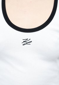 Άσπρο ριγωτό αμάνικο τοπ με μαύρο περίγραμμα γύρω από το ντεκολτέ και μικρό μαύρο κεντητό λογότυπο "KZ" στο κέντρο του στήθους.