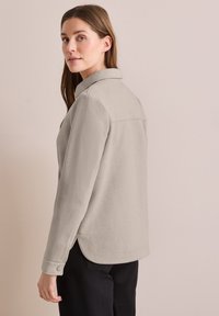 Sudadera texturizada de color gris claro, con collar y puños abotonados, presenta un ligero dobladillo asimétrico y aberturas laterales para mayor comodidad de movimiento.