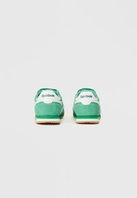 Zalie zamšādas sporta apavi ar baltām akcentiem, kurām ir teksturēta virsma, noapaļota forma un Reebok zīmols uz papēža.