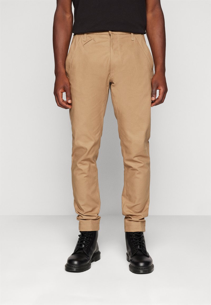 Chinos de color beige con un ajuste slim, elaborados con tejido suave. Presentan bolsillos delanteros y puños enrollados, combinados con botas negras de cordones.