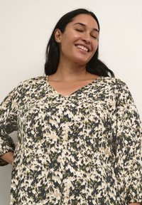 Bluse mit Blumenmuster in Beige, Grün und Dunkelblau. Mit einem V-Ausschnitt, langen Ärmeln und einer lockeren Passform. Leichter Stoff mit weicher Textur.