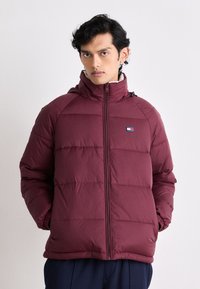 Burgunderfarbene isolierte Jacke mit einem Reißverschluss vorne, hohem Kragen und gestepptem Muster. Verfügt über ein kleines Logo auf der Brust und elastische Bündchen.