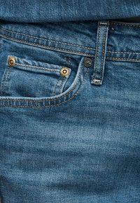 Κοντινό πλάνο τσέπης από μπλε τζιν με εμφανή ραφές, καρφιά και θηλιά ζώνης, φορεμένη με πουκάμισο από τζιν μέσα στην παντελόνα.