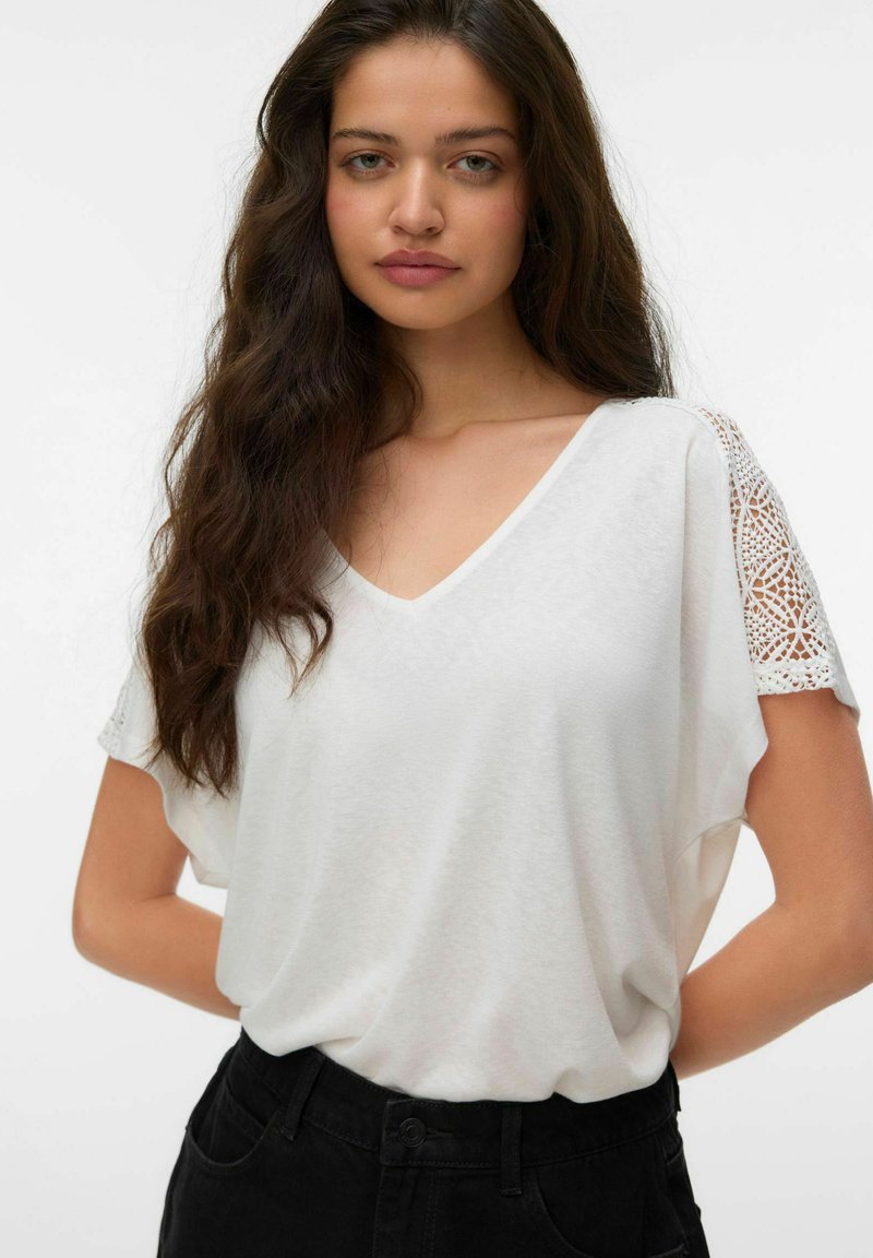 Mujer joven con cabello largo y castaño que lleva una blusa blanca de cuello en V con mangas de encaje y pantalones negros sobre un fondo liso.