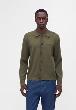 Homme portant un cardigan boutonné vert olive avec col et un jean bleu, debout devant un fond blanc uni.