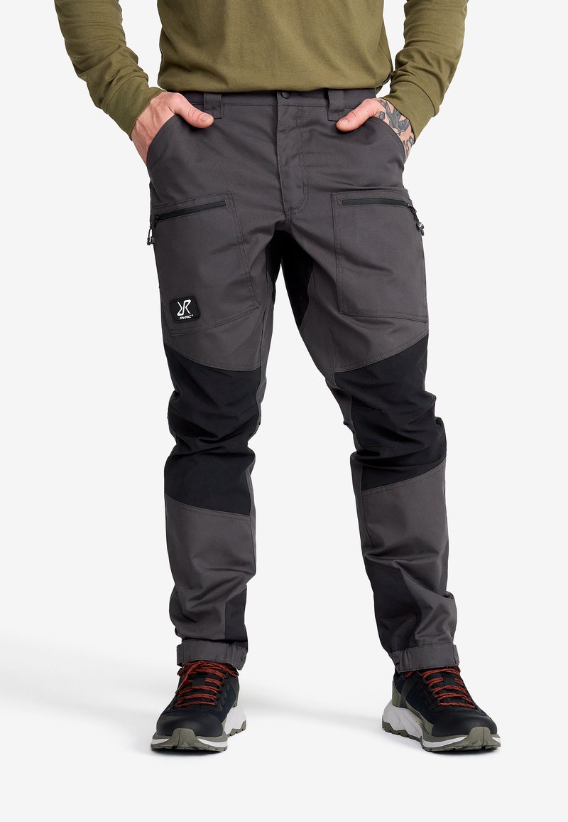 Man som bär grå och svarta cargobyxor med dragkedjefickor på låren, svarta och olivgröna sneakers samt en olivgrön långärmad skjorta, med händerna i fickorna.