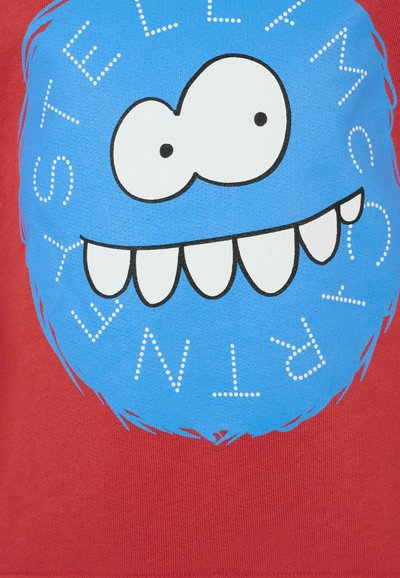 T-shirt rossa con un grande personaggio cartoon blu, rotondo, con occhi e denti bianchi. Il testo attorno al design è in un font giocoso con punti.