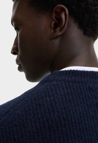 Suéter de punto azul marino con un acabado texturizado, cuello redondo y detalles acanalados; usado por una persona vista de lado.