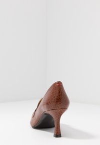 Zapato de tacón alto de charol marrón con un patrón texturizado y embosado, y un tacón negro. Presenta un pequeño acento rojo en el tacón.