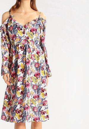 Kobieta ubrana w sukienkę na zimne ramiona, sięgającą do kolan, w różowo-żółto-szary kwiatowy wzór na białym tle.