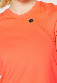 Lähikuva henkilöstä, joka on pukeutunut oranssiin urheiluhaalariin, jossa on tekstuurilla varustetut hartiapaneelit ja pieni musta Under Armour -logo rinnassa.