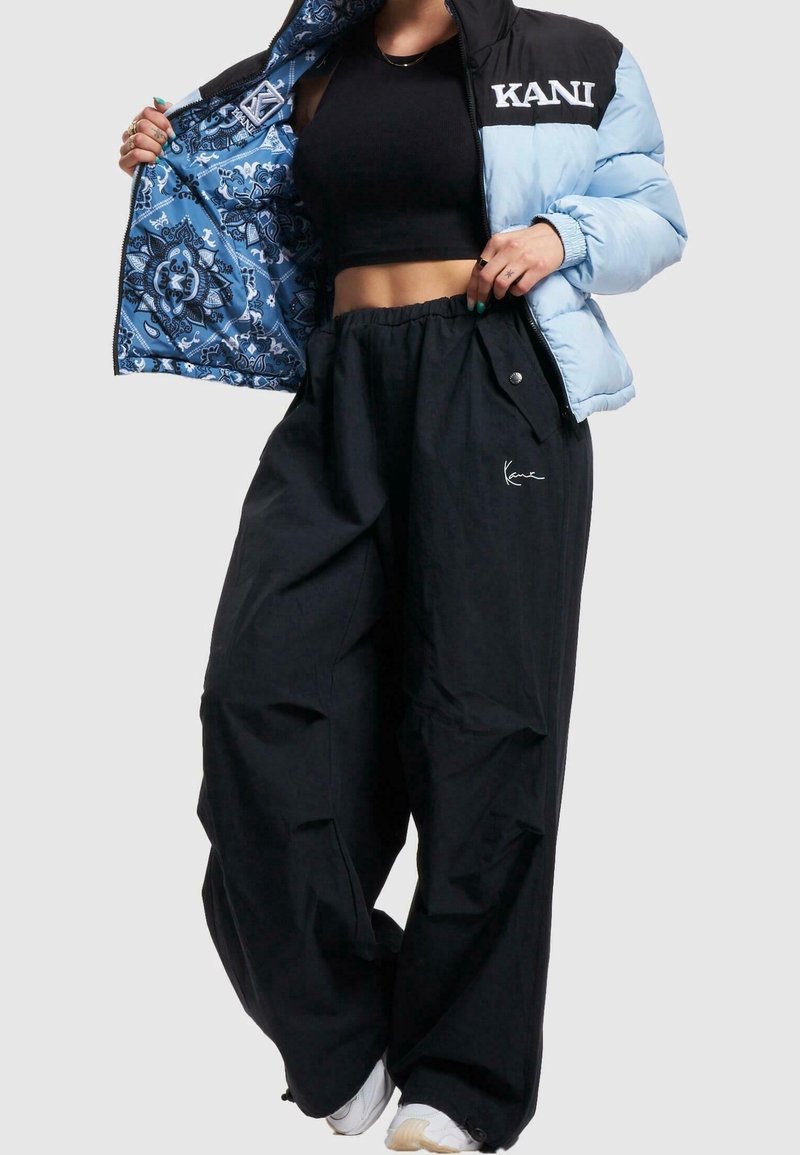 Giacca gonfiabile nera e azzurro chiaro con fodera blu a fantasia e logo, abbinata a pantaloni neri larghi con tasche laterali e dettaglio del logo.