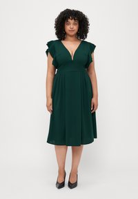 Robe vert foncé avec un décolleté en V profond, des manches courtes à volants et une jupe évasée. Tissu lisse avec des plis, accompagnée de talons hauts noirs.