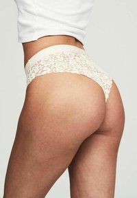 Person som bär vit spetsunderkläder och en vit kort topp, sedd bakifrån mot en enkel ljus bakgrund.