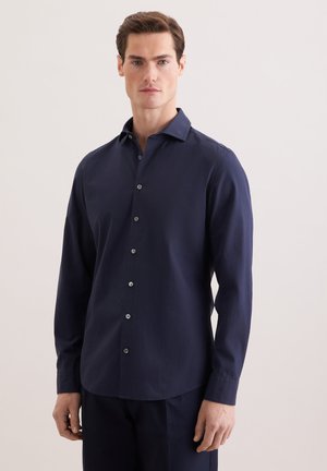 Man die een marineblauw overhemd met lange mouwen draagt, staand tegen een eenvoudige lichte achtergrond.