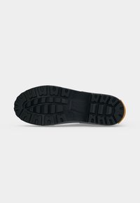 Semelle de chaussure en caoutchouc noir avec des crampons profonds et un motif texturé, présentée sur un fond clair uni.