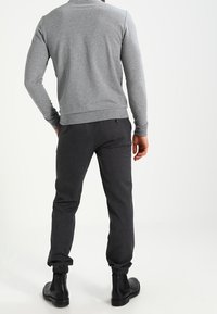Hombre con una camiseta de manga larga gris, pantalones jogger oscuros con puños elásticos y botas negras hasta el tobillo, de pie con las manos en los bolsillos y mirando hacia otro lado.