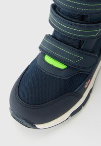 Tuvumā redzama tumši zila sporta apava ar neonzaļiem akcentiem, divām Velcro siksnām, baltu zoli un nelielu karoga detaļu sānos.